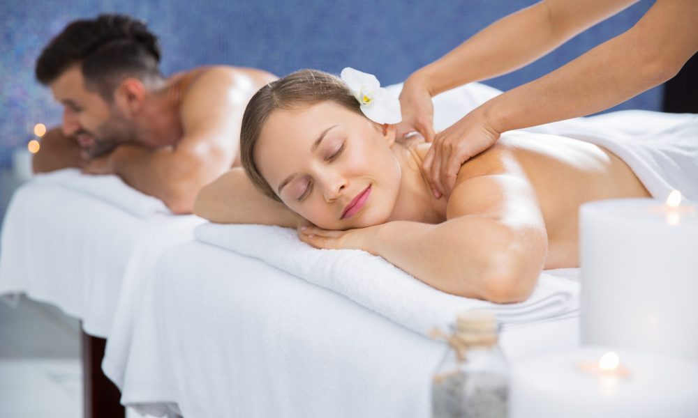 Couple getting massage together in spa center. Young Caucasian woman with flower in hair getting back massage. Man lying on massage table and relaxing after treatment in background
